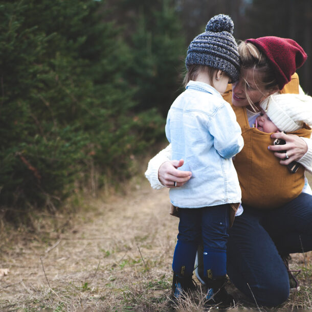 Young mother crouching down to speak to daughter, while holding small baby in carrier on a countryside footpath