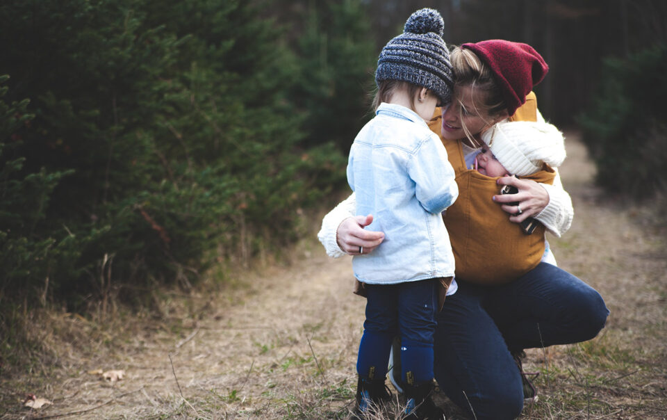 Young mother crouching down to speak to daughter, while holding small baby in carrier on a countryside footpath